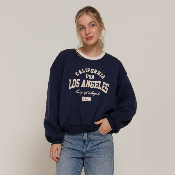 Woman wearing a navy sweatshirt with 'Los Angeles' text and blue jeans on a plain background