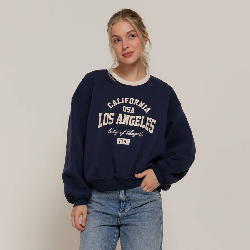 Woman wearing a navy sweatshirt with 'Los Angeles' text and blue jeans on a plain background