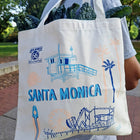 A person outdoors carrying a cream canvas tote bag printed with blue and peach illustrations of a lifeguard tower, palm trees, a Route 66 sign reading Santa Monica, and a boardwalk scene. Kale is visible in the bag's top handles.