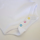 Close-up of a white baby bodysuit or onesie with three colorful snap closures in blue, pink, and yellow on a light beige background.