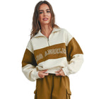 A woman modeling a color-blocked quarter-zip sweatshirt with 'LOS ANGELES' text across the chest, paired with matching brown cargo shorts, white socks, and white sneakers, standing against a plain light gray wall.