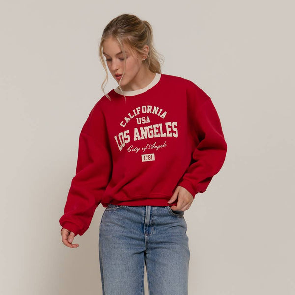Person wearing a red 'Los Angeles' sweatshirt and blue jeans on a plain background