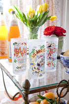 Two colorful tumblers on a glass table with flowers and bottles in the background