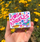 Hand holding a colorful 'Los Angeles' sticker with cartoon illustrations against a yellow flower background.