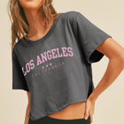 A woman wearing a dark gray cropped t-shirt with 'LOS ANGELES CALIFORNIA' printed in pink and white across the chest, paired with a black skirt, set against a plain beige background.
