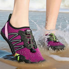 A person standing in shallow ocean water on a beach wearing purple and black water shoes, with one shoe lifted slightly to show the sole and the other sinking into splashing water.