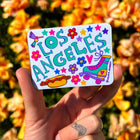 Colorful 'Los Angeles' sticker held in front of a floral background
