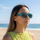 A woman wearing teal-framed sunglasses and a lime green top stands on a sandy beach with the ocean and blue sky in the background.