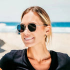 A woman smiling on a sunny beach wearing round black sunglasses and a black top, with a lifeguard tower and ocean in the background.