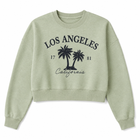 A light sage green cropped crewneck sweatshirt with a black distressed graphic on the front featuring the text 'LOS ANGELES', 'California', palm tree silhouettes, and the numbers '17' and '81'.