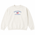 A vanilla crewneck sweatshirt featuring 'SANTA MONICA CALIFORNIA' and a small American flag printed in blue on the chest, displayed against a solid white background.