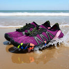 A pair of purple and black water shoes with neon accents, shown on wet sand with ocean waves and a blue sky in the background.