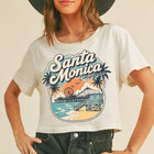A woman wearing a white cropped graphic t-shirt with a colorful 'Santa Monica California' beach print, paired with dark gray jeans and a wide-brimmed dark gray hat, against a solid beige background.