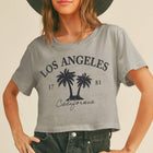 A woman wearing a light gray cropped t-shirt with a black palm tree graphic and 'Los Angeles California' text, paired with a wide-brimmed dark hat and dark denim shorts, against a solid beige background.