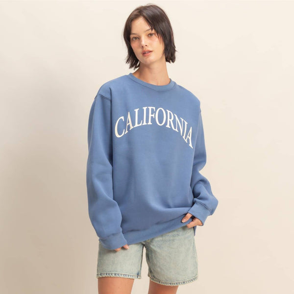 Person wearing a blue 'California' sweatshirt with denim shorts on a beige background. 