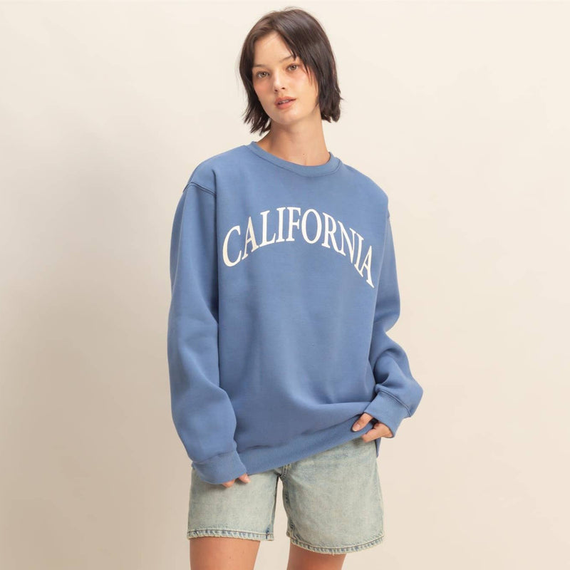 Person wearing a blue 'California' sweatshirt with denim shorts on a beige background. 