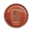 A round brown leather patch with an embossed American flag and the words 'CALIFORNIA' and 'USA', displayed on a rustic wooden background.