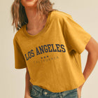 A woman wearing a cropped mustard yellow t-shirt with 'LOS ANGELES CALIFORNIA' printed in navy blue varsity lettering, paired with blue jeans, against a solid beige background.