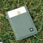 A slim green cardholder wallet with a topographic line pattern, shown lying on green grass with a credit card partially inserted. A small black square logo patch is visible on the lower right.