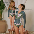 Two people wearing matching floral swimsuits standing indoors with a plant in the background.