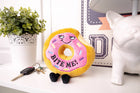 Plush donut toy with 'Bite Me!' text on a surface with keys and a plant in the background