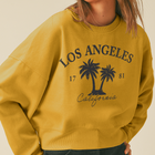 A woman wearing a mustard yellow crewneck sweatshirt with a navy blue 'Los Angeles California' palm tree graphic, paired with dark jeans and a light beige baseball cap, against a solid beige background.