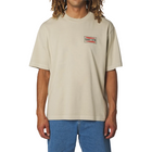 A man wearing a beige short-sleeve t-shirt with a small rectangular chest logo that reads 'DUNES+CAN', paired with blue jeans, against a light gray background.