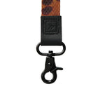 A close-up of a keychain or bag strap featuring a black leather patch with an embossed logo, a black swivel clasp, and a patterned woven strap in orange, brown, and black, set against a plain light gray background.
